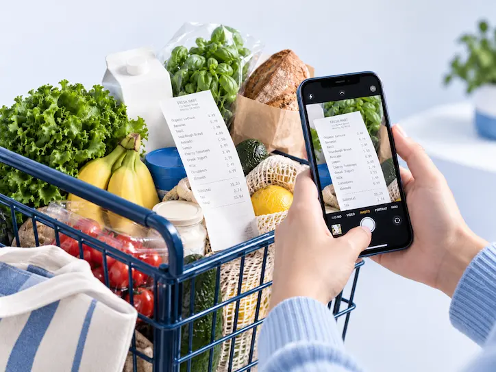 A shopper photographing a grocery receipt beside a cart of groceries.
